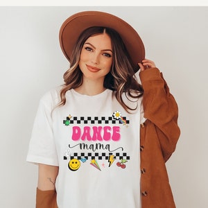 May include: White t-shirt with a black and white checkered background and the text "Dance mama" in pink. The design also includes a variety of colorful cartoon graphics, including a smiley face, a rainbow, a lightning bolt, and a cherry.