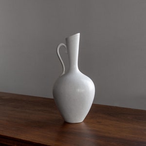 May include: A white ceramic vase with a rounded body and a curved handle. The vase is sitting on a wooden surface.