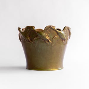 May include: A ceramic planter with a glossy, metallic olive-green glaze. The upper rim features a decorative, scalloped edge with a relief pattern of stylized leaves and berries encircling the top portion of the vessel's exterior surface.