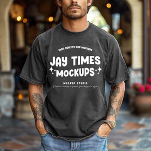 May include: A dark gray t-shirt with the text "JAY TIMES MOCKUPS" in white, with additional text above and below. The shirt is worn by a person in blue jeans.