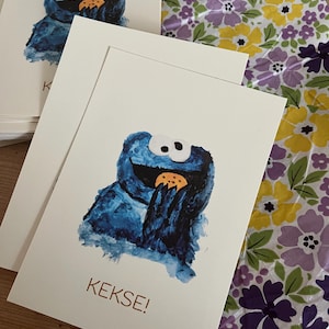 May include: Greeting cards featuring a blue cartoon character eating a cookie. The character has large white eyes and a wide smile. The word "KEKSE!" is printed in brown on the card. A floral patterned fabric is in the background.