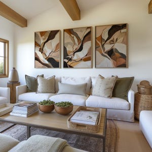 Puede incluir: Un sofá blanco con almohadas verdes, marrones y beige en una sala de estar con tres impresiones de arte abstracto en la pared sobre el sofá. El sofá está frente a una mesa de café con libros y plantas encima.