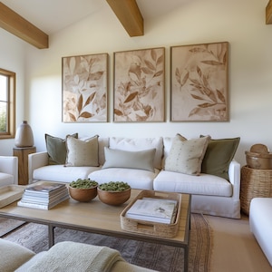 Puede incluir: Un salón con un sofá blanco y cojines en tonos verdes y beige. Tres impresiones botánicas enmarcadas cuelgan sobre el sofá. Una mesa de centro de madera exhibe libros, cuencos de madera y una bandeja tejida, creando una paleta de colores neutros.