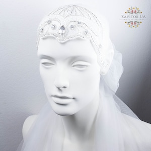 May include: A white beaded headband with a large crystal in the center. The headband is on a white mannequin head with a white veil.