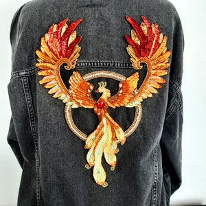 May include: A denim jacket with a large embroidered phoenix patch. The phoenix is made of sequins and beads in shades of red, orange, and gold. The phoenix is surrounded by a circle of sequins.