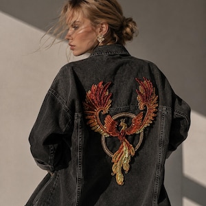 Könnte beinhalten: Dunkelgraue Jeansjacke mit einem großen, farbenfrohen Phönix-Design, das auf den Rücken gestickt ist. Der Phönix hat rote, orangefarbene und goldene Pailletten. Die Jacke hat ein klassisches Knopfdesign.
