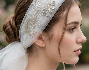 May include: A white tulle headband featuring intricate silver embroidery and clear crystal embellishments in a swirling floral pattern. The sheer fabric band wraps around the head, with a soft tulle veil extending from the back.
