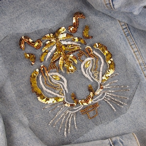 Peut inclure: Un visage de tigre brodé de sequins dorés et blancs sur une veste en jean bleue.