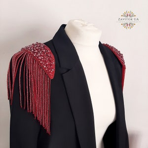 Op de afbeelding: Een zwart blazer met rode kralenversieringen op de schouders. De versieringen zijn gemaakt van kleine, ronde kralen en hebben een franje-achtig ontwerp.
