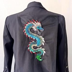 Könnte beinhalten: Schwarzer Blazer mit einem farbenfrohen Drachendesign auf dem Rücken. Der Drache ist mit blauen, grünen, roten und silbernen Pailletten bestickt. Der Blazer hat lange Ärmel und eine klassische Passform.