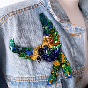 May include: A blue denim jacket with a colorful embroidered bird applique. The bird is made of sequins and beads in shades of blue, green, yellow, and gold.