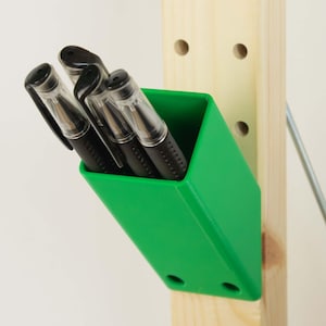 Könnte beinhalten: Ein leuchtend grüner Stifthalter mit vier schwarzen Stiften. Der Halter ist an einem hellen Holzpfosten befestigt. Die Stifte haben transparente Schäfte und schwarze Kappen.