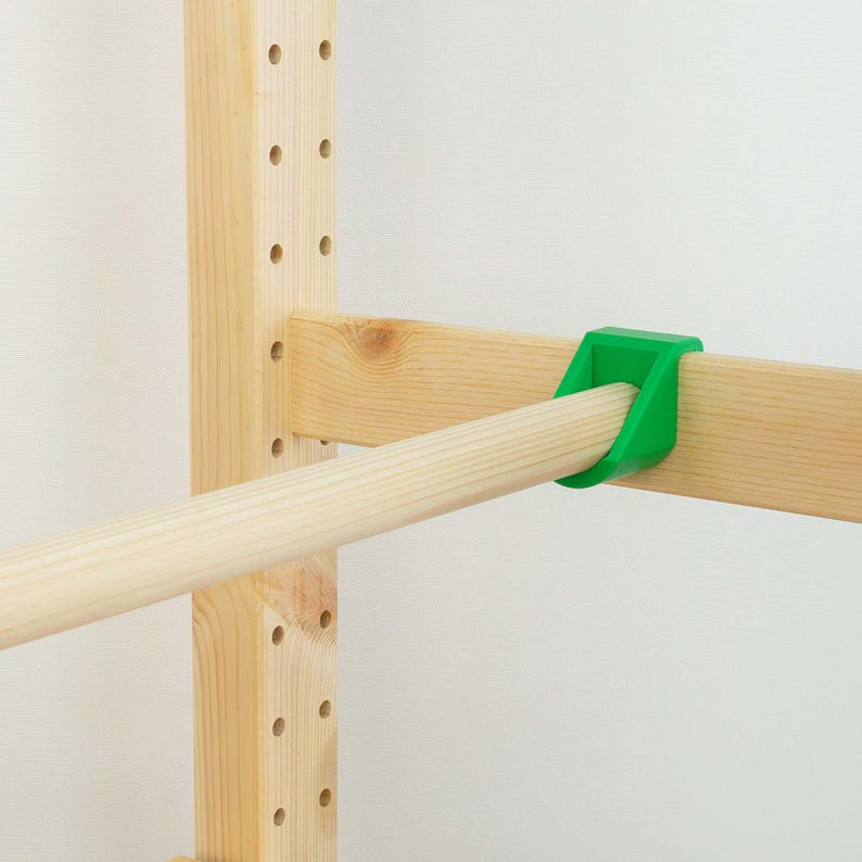 May include: A wooden climbing frame with a green plastic connector. The frame is made of light-colored wood and features a horizontal bar held in place by the green connector. The background is a plain white wall.