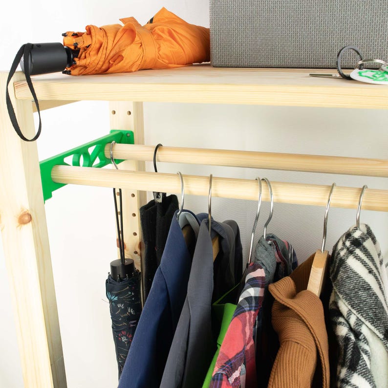 May include: A wooden clothing rack with a top shelf holding an orange umbrella and a gray box. Below, a wooden rod holds various garments on hangers. A green bracket supports the rod. The rack is against a white wall.