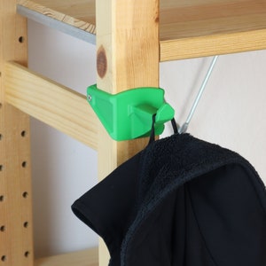 Può includere: Un gancio angolare verde brillante fissato a un supporto per scaffale in legno. Una giacca nera con interno sfocato è appesa al gancio. Lo scaffale è in legno chiaro.