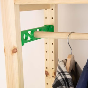 Può includere: Organizzatore per armadio in legno con una staffa di supporto verde e un'asta di legno per appendere i vestiti. Una gruccia di legno tiene una giacca a quadri bianchi e neri. La struttura dell'armadio è in legno chiaro.