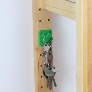 Könnte beinhalten: Ein grüner magnetischer Haken, der an einer Holz-Lochwand befestigt ist, mit einem Schlüsselbund, der daran hängt.