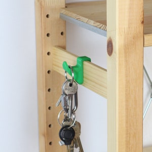 Könnte beinhalten: Ein grüner Plastikhaken, der an einem Holzregal befestigt ist, an dem ein Schlüsselbund hängt. Die Schlüssel sind silberfarben und haben einen schwarzen Schlüsselanhänger.