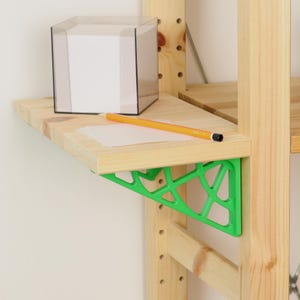 Può includere: Una staffa per mensola triangolare traforata di colore verde brillante fissata a uno scaffale in legno chiaro. Sul ripiano in legno si trova un cubo in plastica trasparente contenente un blocco di fogli bianchi e una matita gialla sopra.