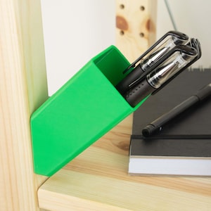 Könnte beinhalten: Ein leuchtend grüner Stifthalter, schräg auf einem Holzregal, enthält zwei schwarze Stifte mit transparenten Gehäusen. Ein schwarzer Stift und ein Notizbuch sind ebenfalls vorhanden. Ein Büroaccessoire.
