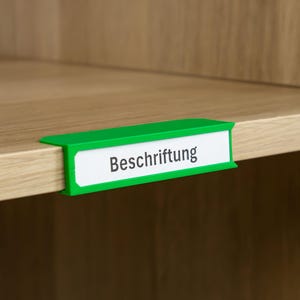 Puede incluir: Un portaetiquetas verde con la palabra "Beschriftung" impresa en una etiqueta blanca. El portaetiquetas está fijado al borde de una estantería de madera.
