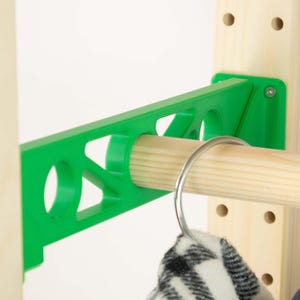 May include: A green plastic clothing rack support with a wooden dowel and a metal hanger. The support is attached to a light-colored wooden frame. A black and white patterned garment hangs from the hanger.