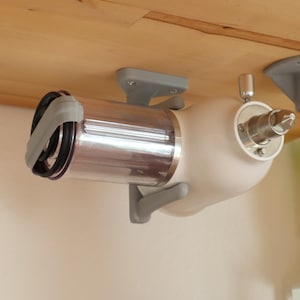 Op de afbeelding: Een wit en zilver keukenapparaat, mogelijk een vleesmolen, gemonteerd onder een houten oppervlak. Het apparaat heeft een cilindrische zilveren component en een witte behuizing, bevestigd met grijze beugels.