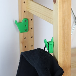 Può includere: Due ganci di plastica verde montati su uno scaffale di legno. Una giacca di pile nera è appesa a uno dei ganci.