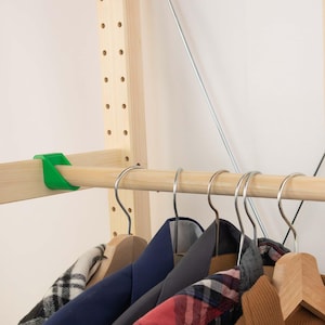 Può includere: Barra appendiabiti in legno con grucce in metallo che contengono vari capi. Un supporto in plastica verde fissa la barra al telaio in legno. Il telaio presenta fori distanziati uniformemente. L'abbigliamento comprende una camicia a quadri, una giacca blu navy e una giacca di velluto a coste marrone.