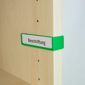 Può includere: Un portaetichette verde con la parola "Beschriftung" in testo nero, attaccato a una superficie di legno di colore chiaro. Il portaetichette è rettangolare con un inserto bianco.