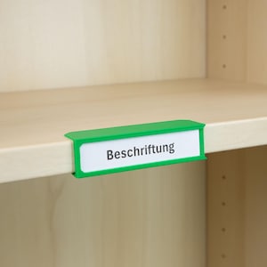 Può includere: Un portaetichette verde con la parola "Beschriftung" stampata su un'etichetta bianca. Il portaetichette è attaccato a una mensola di legno. Lo sfondo è un armadio di legno colorato chiaro.
