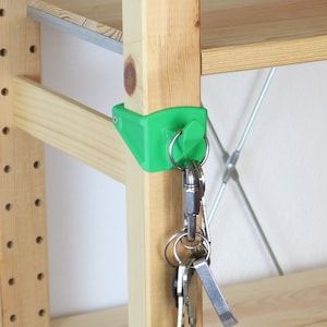 Può includere: Un portachiavi verde stampato in 3D attaccato a uno scaffale di legno. Il portachiavi ha un anello per le chiavi e un piccolo gancio. Un set di chiavi è appeso all'anello.