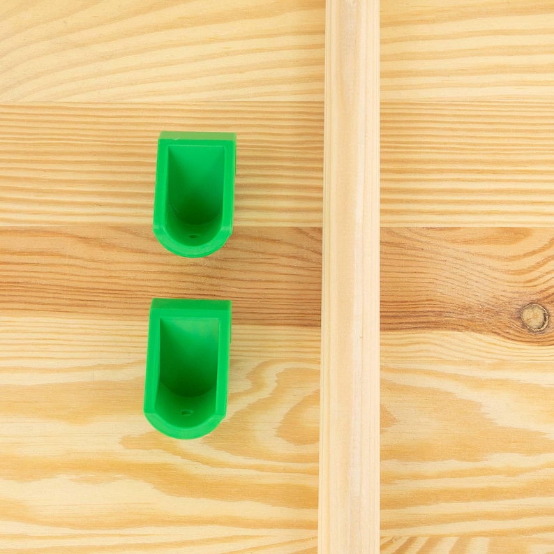 May include: Two bright green, U-shaped plastic holders and a light-colored wooden dowel are set against a light-colored wood grain background. The holders are positioned vertically, and the dowel runs parallel to them.