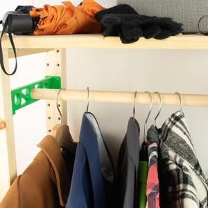 May include: A wooden clothing rack with a green support bracket and a wooden rod for hanging clothes. Various garments hang on metal hangers, including a brown coat, blue shirts, and a plaid jacket. An orange umbrella and black gloves rest on the top shelf.