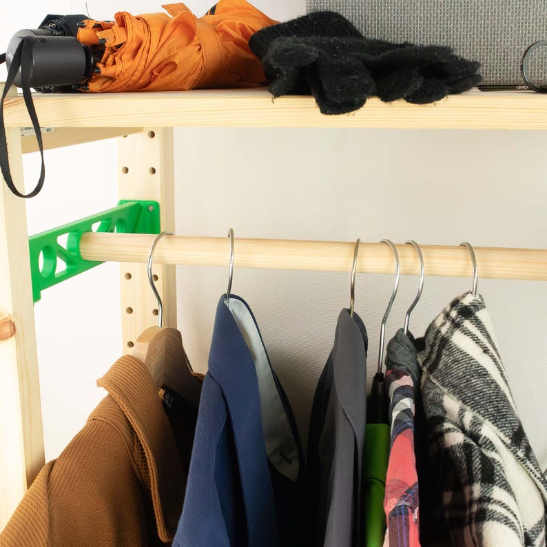 May include: A wooden clothing rack with a green support bracket and a wooden rod for hanging clothes. Various garments hang on metal hangers, including a brown coat, blue shirts, and a plaid jacket. An orange umbrella and black gloves rest on the top shelf.