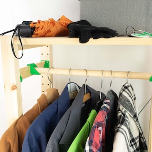 May include: A wooden clothing rack with a top shelf holding an orange umbrella, black gloves, and keys. The lower level has a wooden rod with hangers holding various jackets and shirts in shades of brown, blue, green, red, and black.