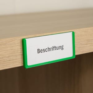Może przedstawiać: Biała etykieta z napisem "Beschriftung" w czarnym tekście, obramowana zieloną obwódką, przymocowana do jasnej drewnianej powierzchni. Etykieta jest prostokątna i wygląda na znak lub znacznik.