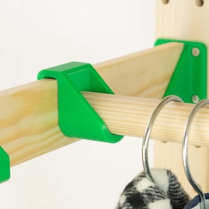 May include: Close-up of a wooden clothing rack with green plastic supports. The rack has a natural wood finish and holds a metal rod for hanging clothes. A black and white patterned garment hangs from the rod.