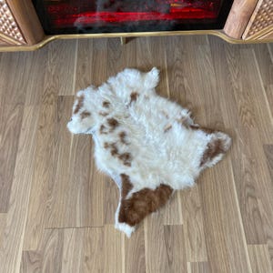 May include: A sheepskin rug with a natural pattern of brown spots and patches on a white background. The rug is irregularly shaped and has a soft, fluffy texture. It is placed on a wooden floor.