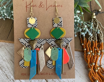May include: Dangle earrings featuring layered geometric shapes in black and white patterns, solid green, blue, orange, and metallic gold. The earrings include small gold-tone metal hearts and rectangular bars, suspended from gold-tone metal hooks.