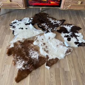 May include: A collection of six cowhide rugs in various colors and patterns. The rugs feature a mix of brown, white, and black spots and patches. The rugs are arranged on a light-colored wooden floor.