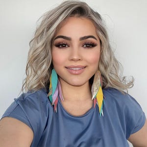 Peut inclure: Une femme aux cheveux blonds ondulés portant une chemise bleue et de longues boucles d'oreilles en plumes colorées. Les boucles d'oreilles sont en cuir et ont un schéma de couleurs or, turquoise, rose et jaune.