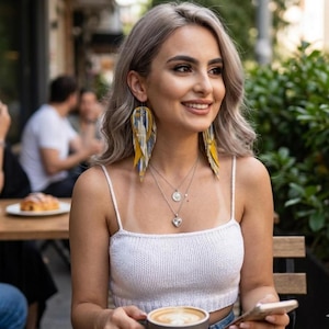May include: A woman wearing long, yellow, blue, and white feather earrings. She is wearing a white knit top and multiple silver necklaces. She is holding a cup of coffee and a phone. The background is blurred, suggesting an outdoor cafe setting.