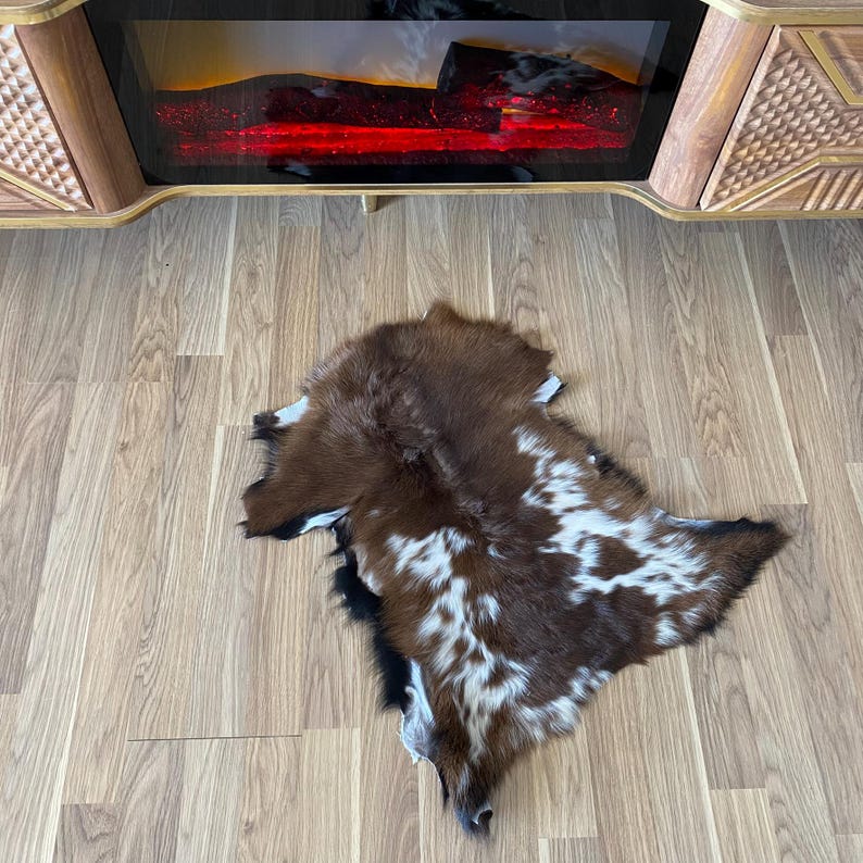 May include: A brown and white cowhide rug with a natural shape. The rug has a mix of brown, white, and black fur. The rug is lying on a light-colored wooden floor.