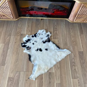 May include: A white and black cowhide rug with irregular edges. The rug has a spotted pattern of black spots on a white background. The rug is lying on a wooden floor.