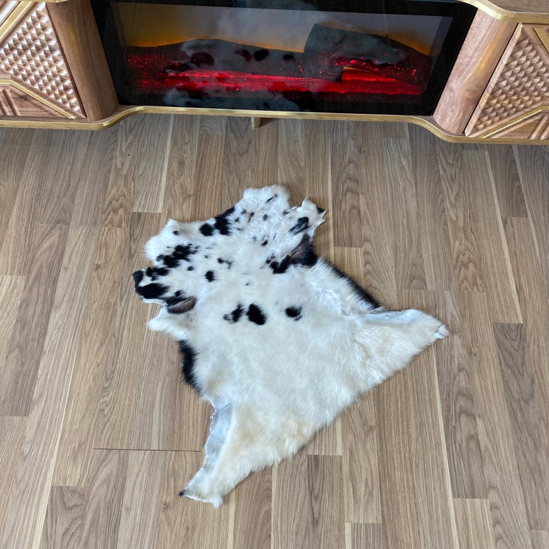 May include: A white and black cowhide rug with irregular edges. The rug has a spotted pattern of black spots on a white background. The rug is lying on a wooden floor.
