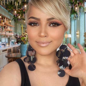 May include: A woman with short blonde hair wearing large black floral earrings. The earrings are made of fabric and have a layered design.