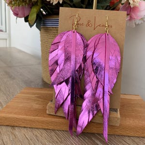 May include: A pair of long, pink metallic feather earrings. The earrings are made of layered leather and have a shiny finish. The earrings are hanging from gold hooks.