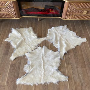 May include: Three off-white, fluffy sheepskin rugs are arranged on a wooden floor. The rugs have irregular shapes and a soft, textured appearance. The background includes a wooden cabinet with a fireplace.