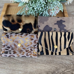 Puede incluir: Cuatro pequeñas carteras con estampados de animales. Las carteras incluyen leopardo, camuflaje, cebra y un patrón dorado. Cada cartera tiene un cierre de botón a presión y se exhiben en una bandeja de madera.